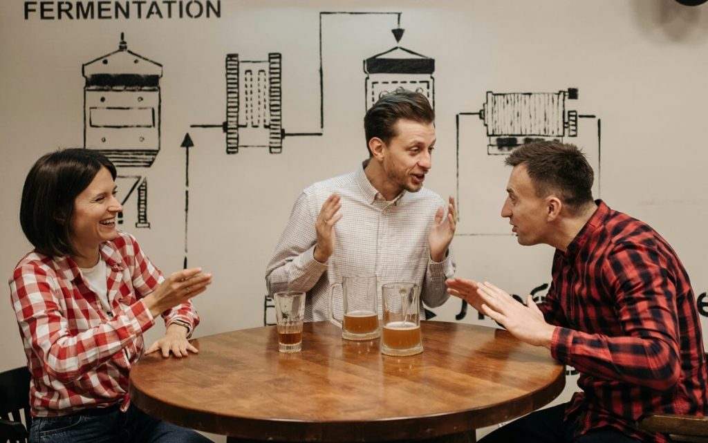Links eine Frau, rechts neben ihr zwei Männer, die um einen Tisch herum sitzen auf dem Biergläser stehen