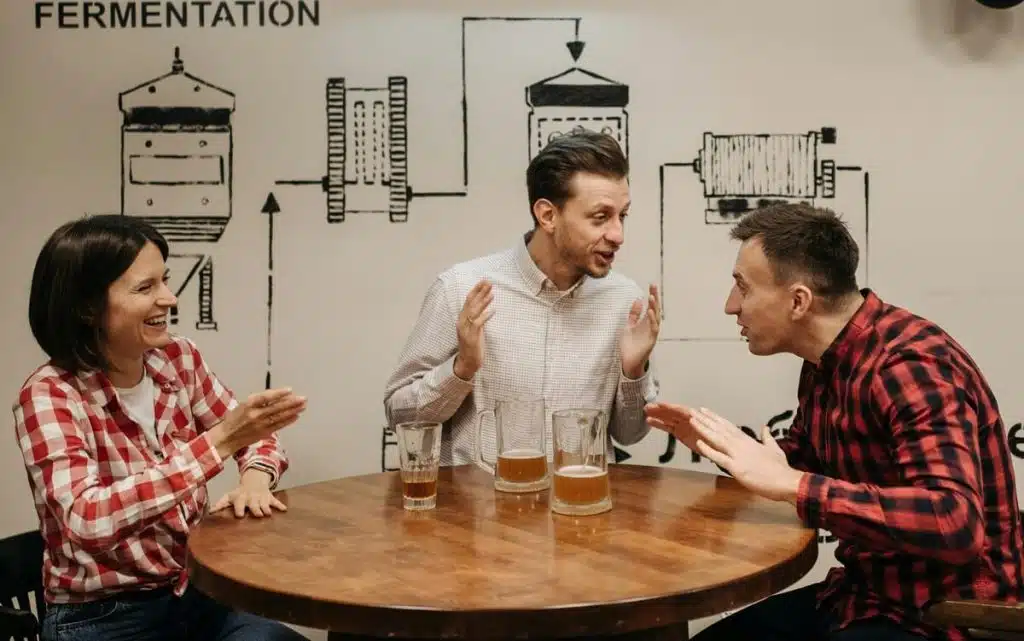 Links eine Frau, rechts neben ihr zwei Männer, die um einen Tisch herum sitzen auf dem Biergläser stehen