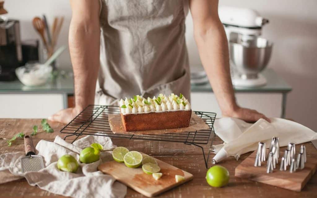 Fertig gebackener Kastenkuchen auf Kuchengitter, dekoriert mit Limettenscheiben. Im Hintergrund eine Person mit Schürze