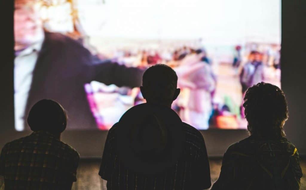 Im Hintergrund läuft ein Film auf einer Leinwand, vorne sind schemenhaft Personen zu sehen.