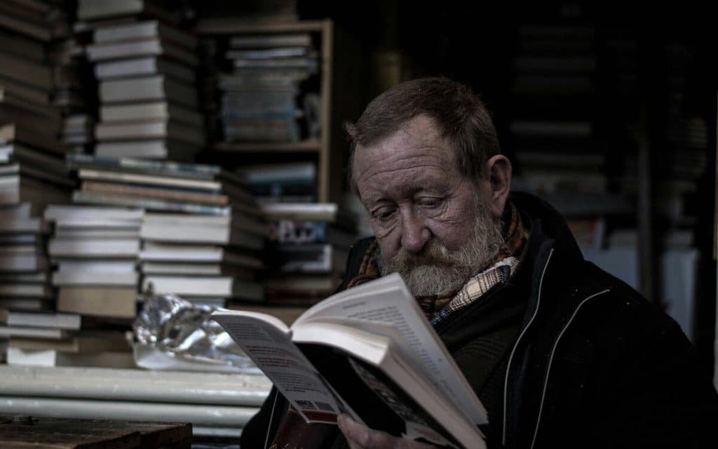 Älterer Mann mit einem Buch in der Hand, im Hintergrund Bücherstapel