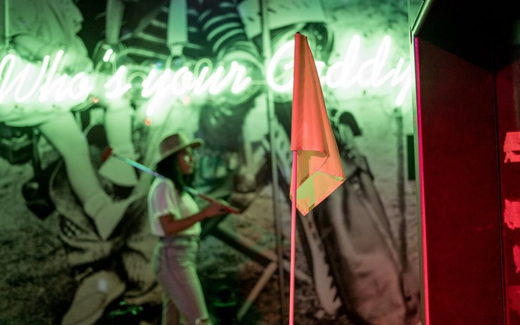 Orangene Golffahne, im Hintergund Neonlicht und eine Person mit Minigolfschläger