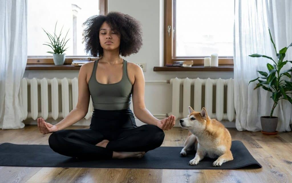 Frau sitzt im Schneiderseitz und mach Yoga, ein Corgi sitzt rechts neben ihr